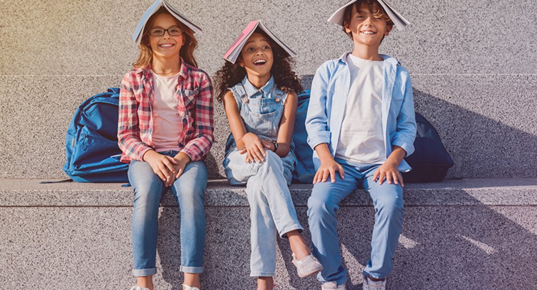 Drei fröhliche Schulkinder sitzend auf Stufe mit Schulbüchern auf dem Kopf