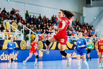 Handballspiel in einer Sporthalle: Eine Spielerin im roten Trikot springt mit Ball zum Torwurf, während mehrere Gegenspielerinnen in blauen Trikots verteidigen. Im Hintergrund verfolgen zahlreiche Zuschauerinnen und Zuschauer das Spiel von der Tribüne aus.