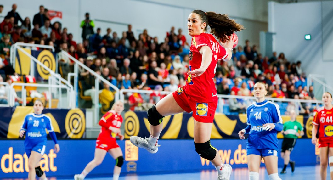Handballspiel in einer Sporthalle: Eine Spielerin im roten Trikot springt mit Ball zum Torwurf, während mehrere Gegenspielerinnen in blauen Trikots verteidigen. Im Hintergrund verfolgen zahlreiche Zuschauerinnen und Zuschauer das Spiel von der Tribüne aus.