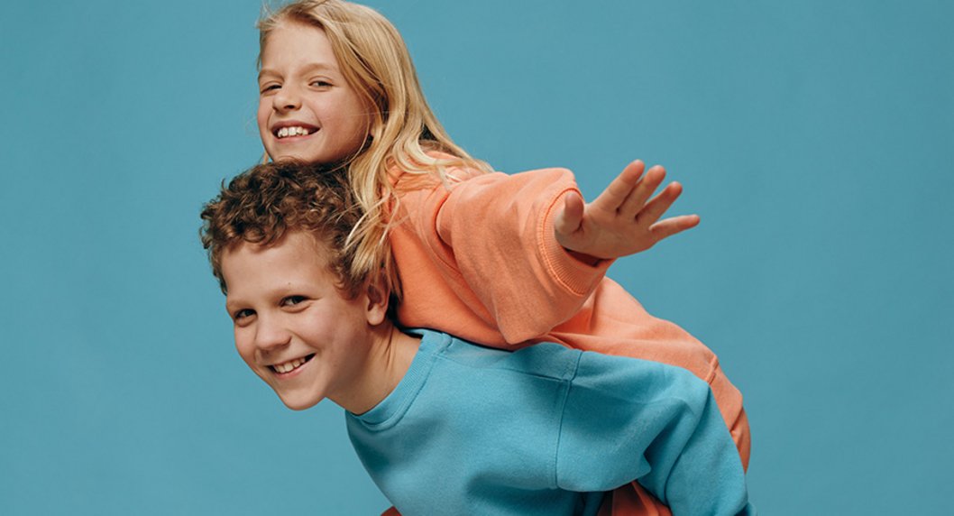 Lachender Junge und lachendes Mädchen in bequemer Kleidung auf hellblauem Hintergrund, Junge trägt Mädchen huckpack